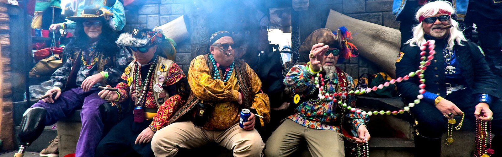 Cigar smoking pirates enjoy the ride and toss beads from their float, surrounded by the smoke of the guns fired by the krewe captain above.