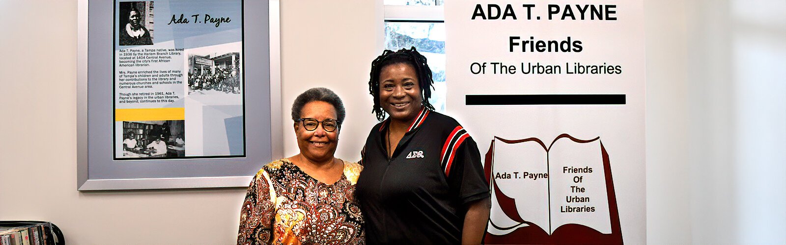 Mary M. James, President of Ada T. Payne Friends of the Urban Libraries, and volunteer Darlene Harris pose in front of a write-up on Ada T. Payne, Tampa’s first African American librarian and Mary’s grandmother.