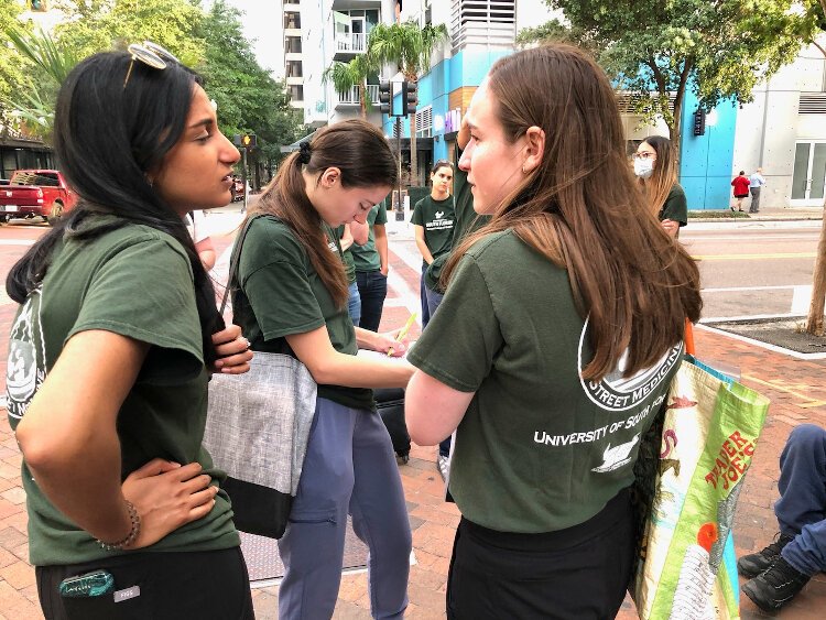 Morsani College of Medicine students Apoorva Ravichandran, Allison O. Dumitriu Carcoana and Lila Gutstein stand by as some students tend to a homeless man on the sidewalk. Ravichandran and Gutstein are co-presidents of Tampa Bay Street Medicine.