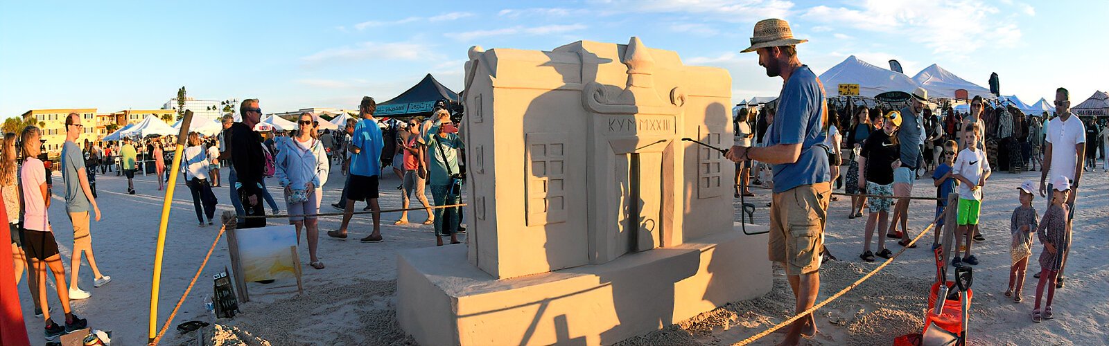 Festival goers watch French sculptor Benoit Dutherage apply a coat of wood glue-based sealant to further protect the “Rock the Block” sculpture he created with other artists.
